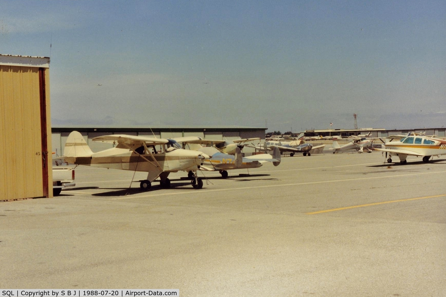 San Carlos Airport (SQL) - N2371P at San Carlos in 1988.View is  north and the administration building (flag) and  Sky Kitchen. Notice two airliners above 71P on (short) final to SFO.Was interesting flying under them (tower gave advisorys) when departing to the east.