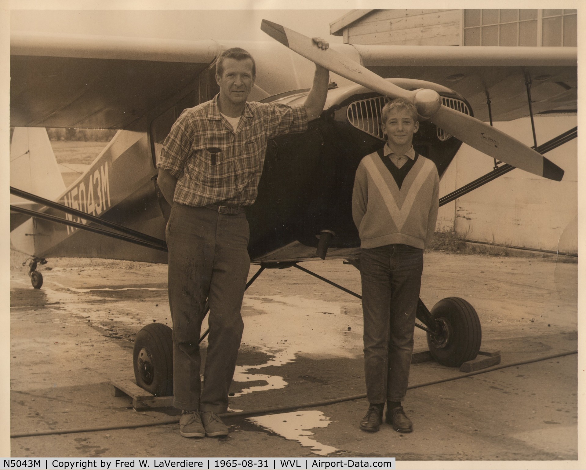 N5043M, 1946 Taylorcraft BC-12D-85 Sportsman C/N 10343, (Left-to-right:) Gerald L. Robbins, Stephen L. Robbins. Stamped on back: “Date AUG 31 1965; Neg. no. 1; Photo by Fred W. LaVerdiere, Waterville, Maine”. With Gerry’s 1946 Taylorcraft at Waterville Robert Lafleur Airport in Waterville, Maine.